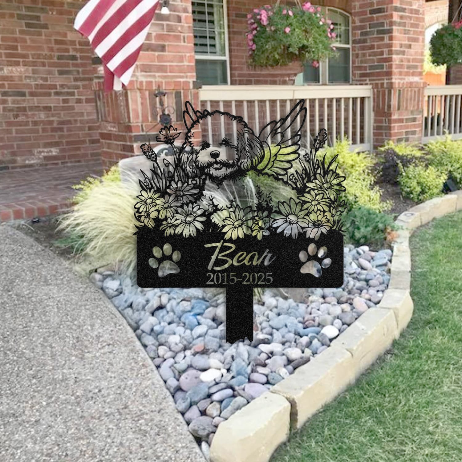 Bichon Frise pet grave marker placed in a rock garden bed in front of a brick house porch with an American flag