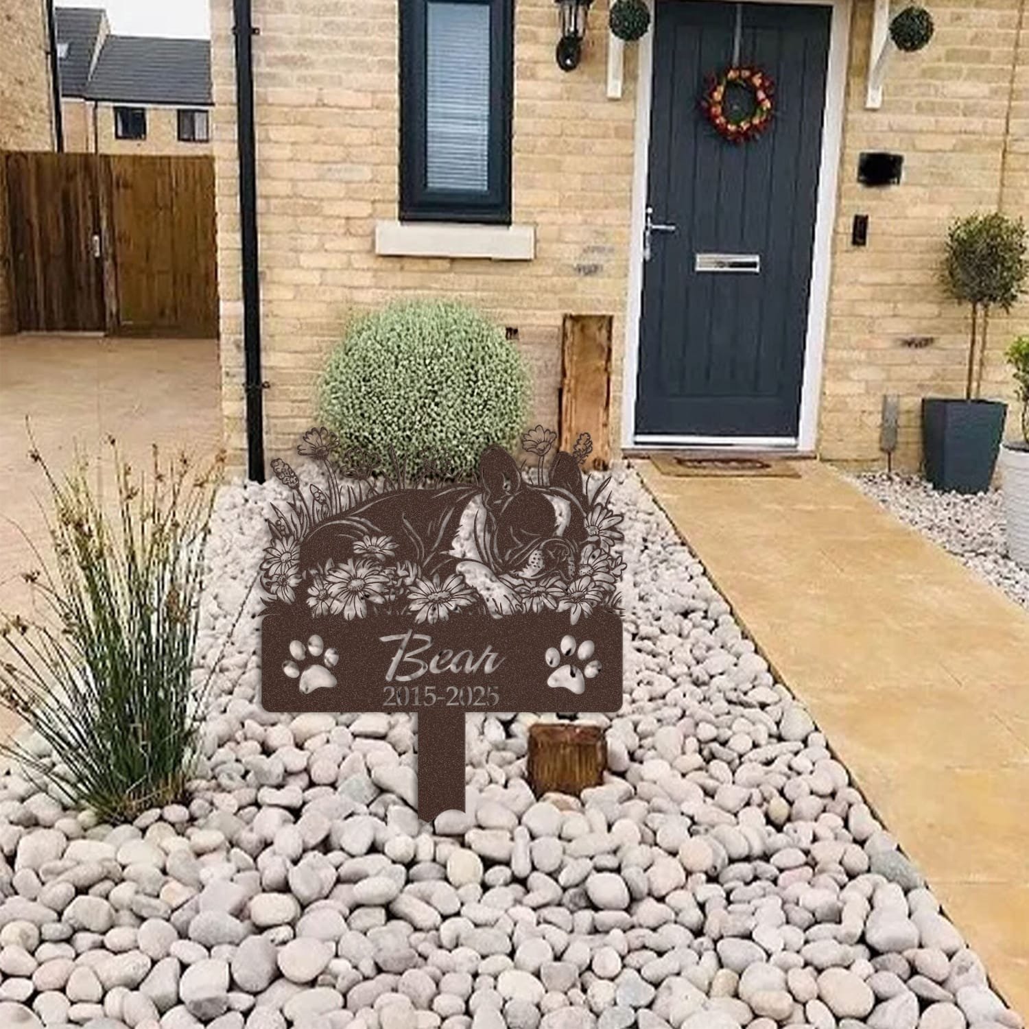 Boston Terrier pet grave marker in a light stone garden bed outside a brick home with a blue front door and wreath