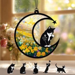 Crescent moon suncatcher with a black-and-white Tuxedo Cat silhouette and raised paw over a stained-glass daffodil field