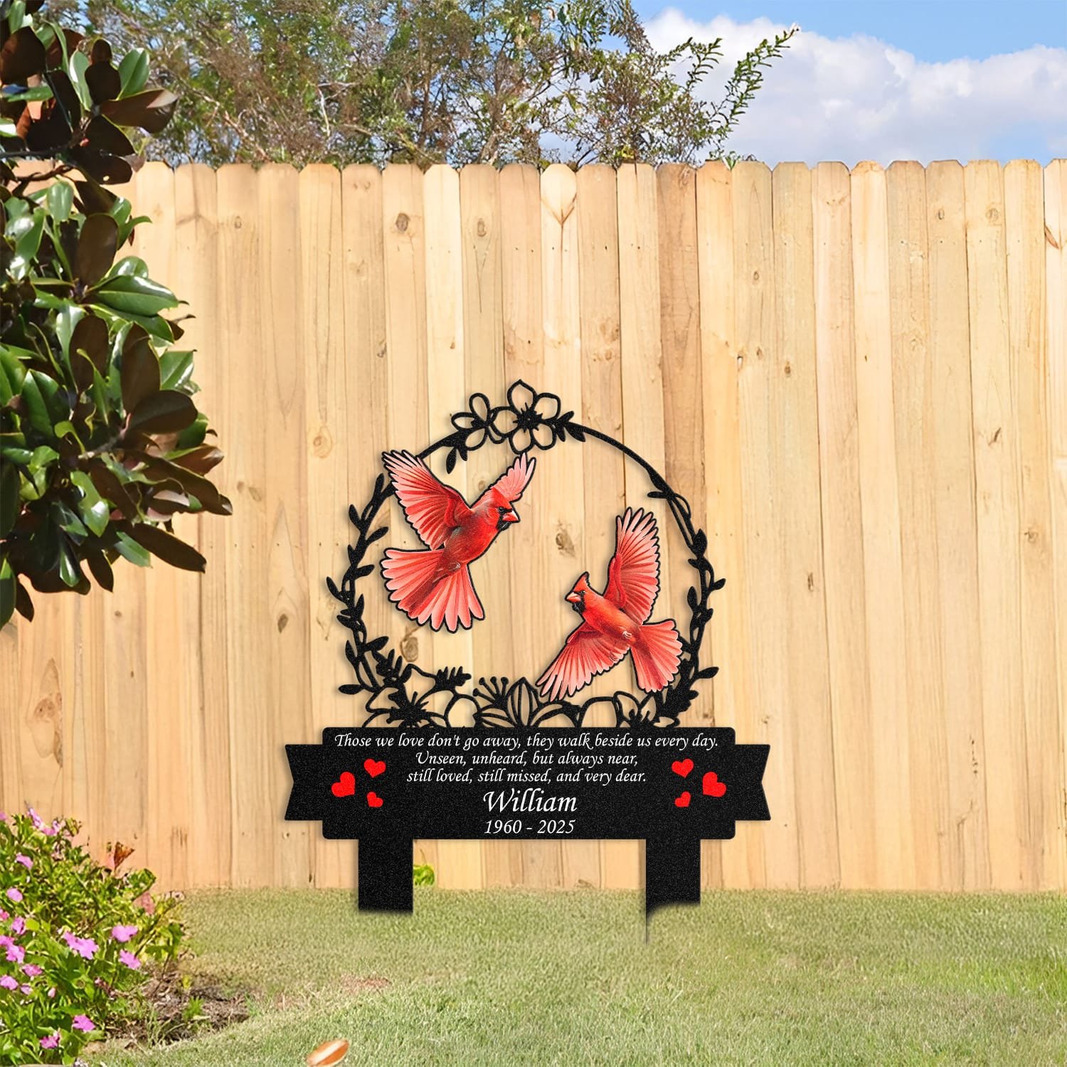 Cardinal human grave marker displayed among garden plants and stone edging, showing the floral wreath bird-pair design as an outdoor memorial accent.
