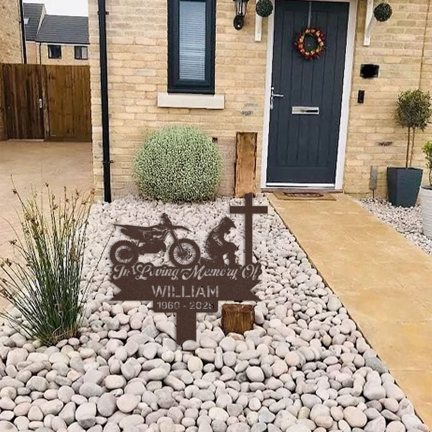Dirt Bike human grave marker displayed among garden plants and stone edging, showing the dirt bike kneeling-at-the-cross design as an outdoor memorial accent.