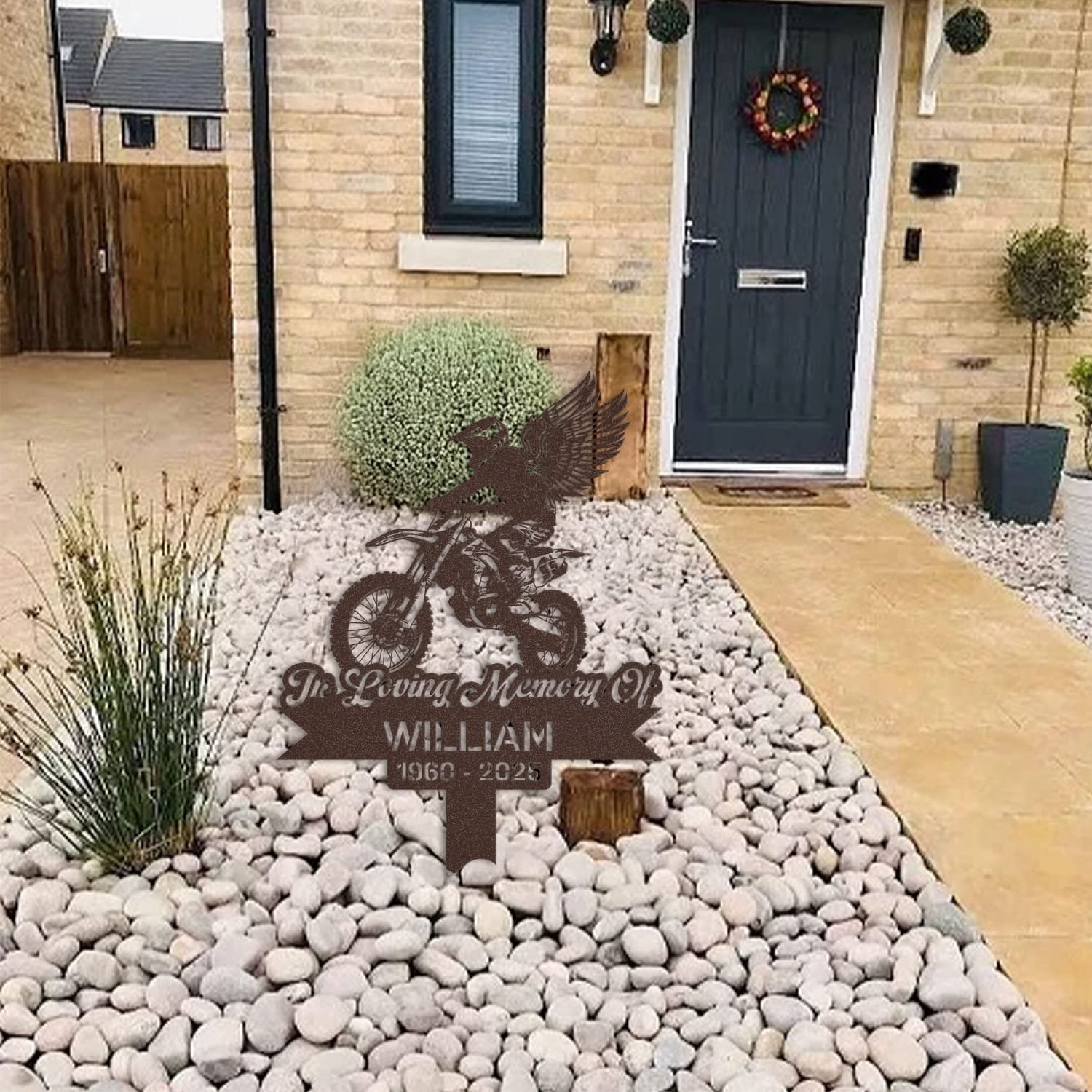 Dirt Bike human grave marker displayed among garden plants and stone edging, showing the dirt bike winged rider design as an outdoor memorial accent.