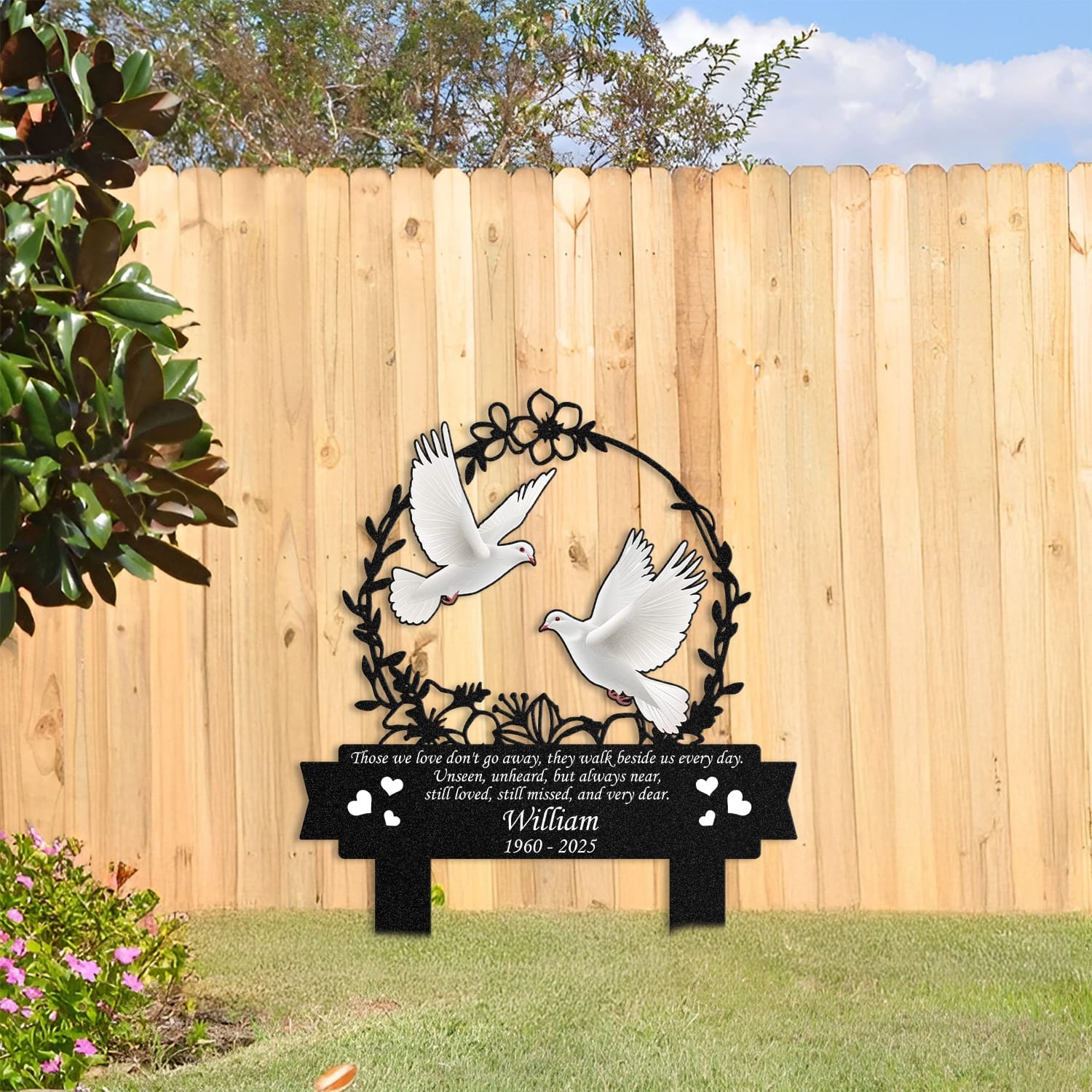 Dove human grave marker displayed among garden plants and stone edging, showing the floral wreath bird-pair design as an outdoor memorial accent.