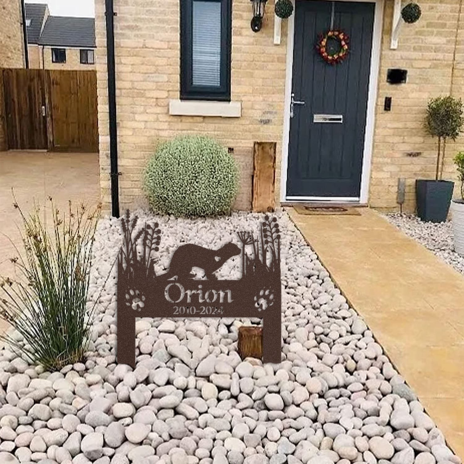 Ferret pet grave marker displayed among garden plants and stone edging, showing the heart-cutout silhouette design as an outdoor memorial accent.
