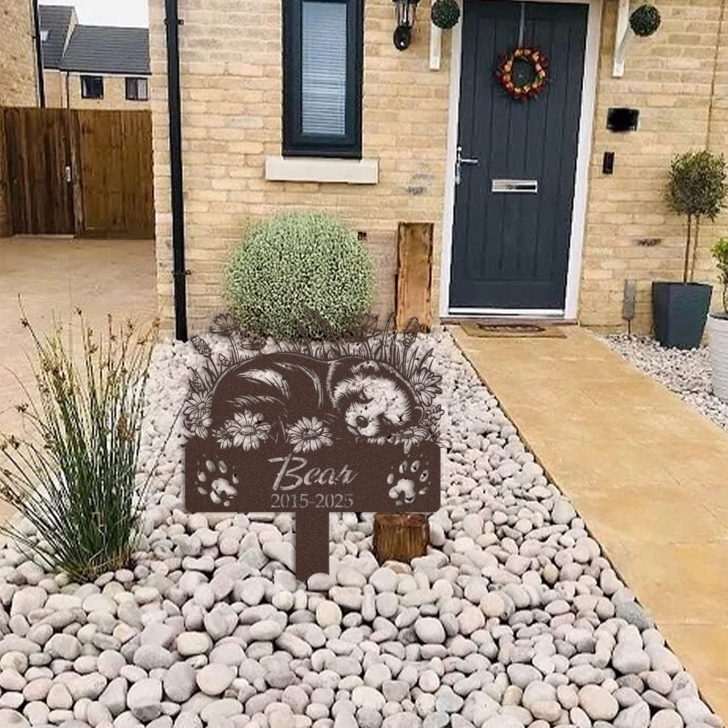 Ferret pet grave marker displayed among garden plants and stone edging, showing the sleeping-pose daisy floral design as an outdoor memorial accent.