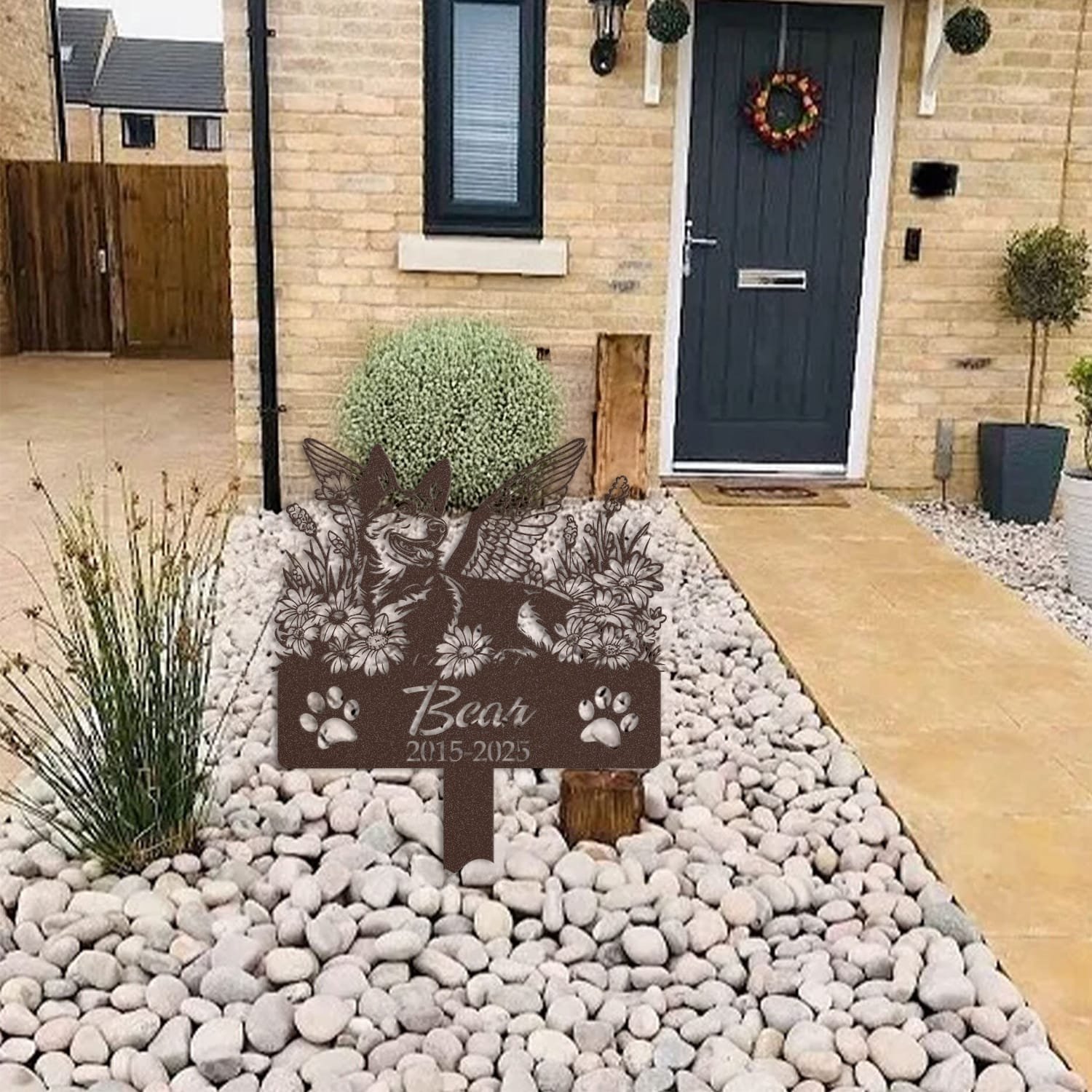 German Shepherd pet grave marker displayed among garden plants and stone edging, showing the angel wings and daisy floral design as an outdoor memorial accent.