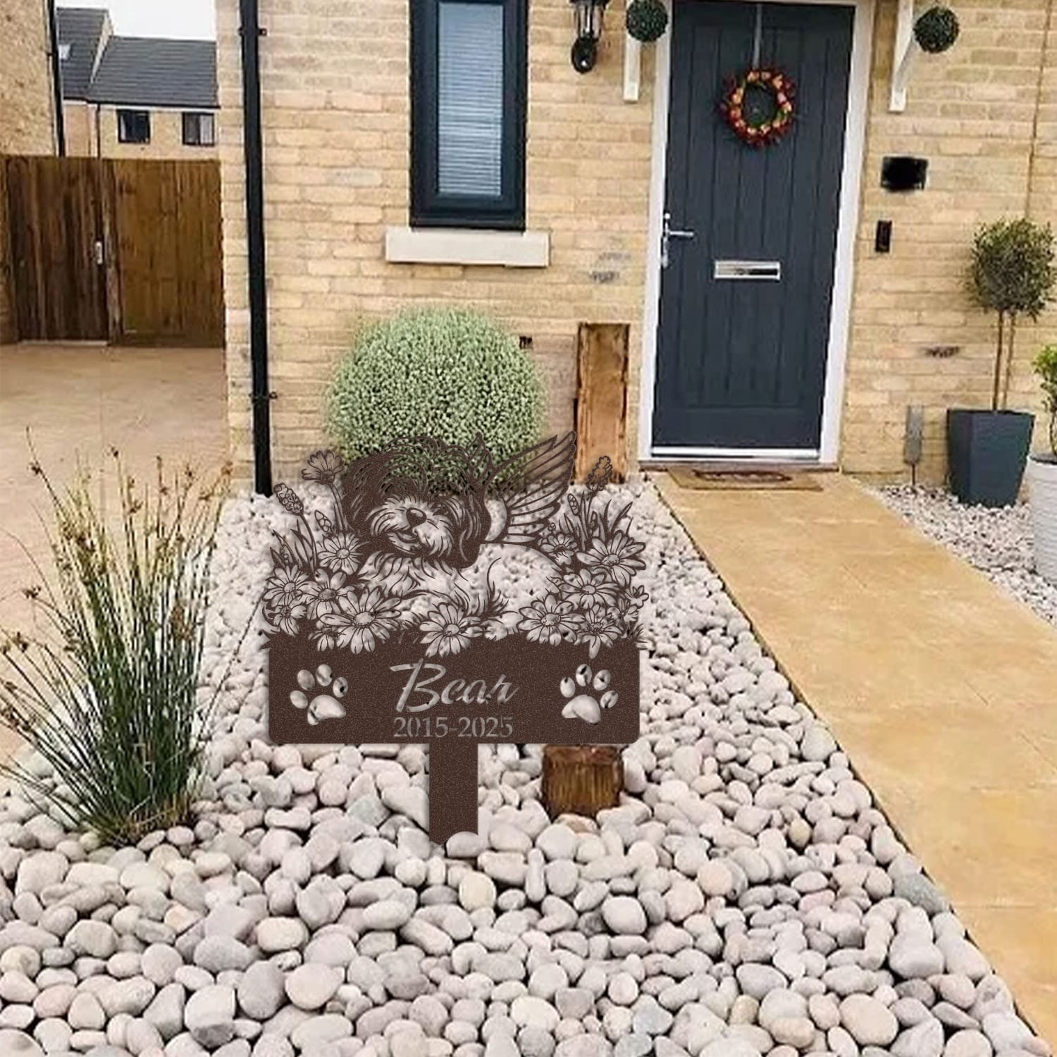 Havanese pet grave marker displayed among garden plants and stone edging, showing the sleeping-pose daisy floral design as an outdoor memorial accent.