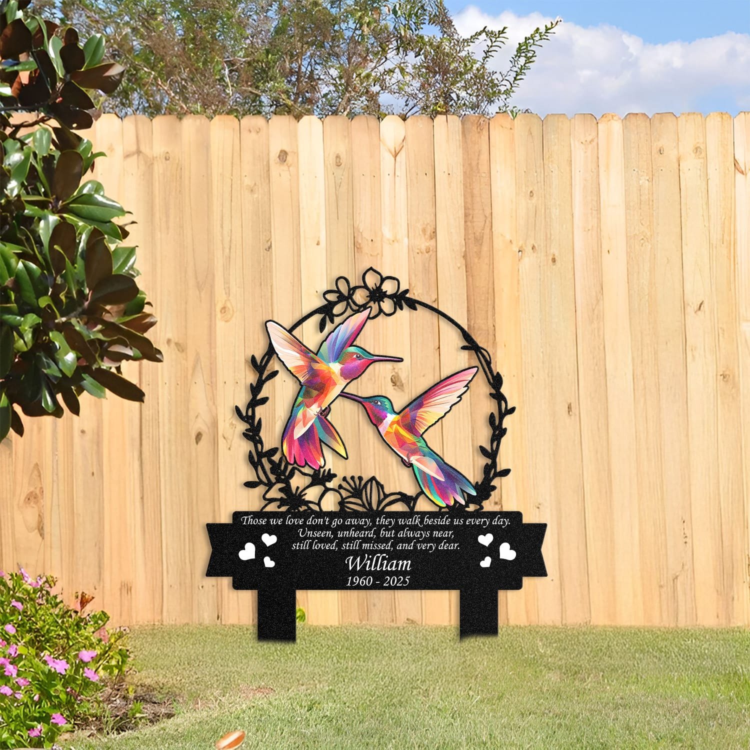 Hummingbird human grave marker displayed among garden plants and stone edging, showing the floral wreath bird-pair design as an outdoor memorial accent.