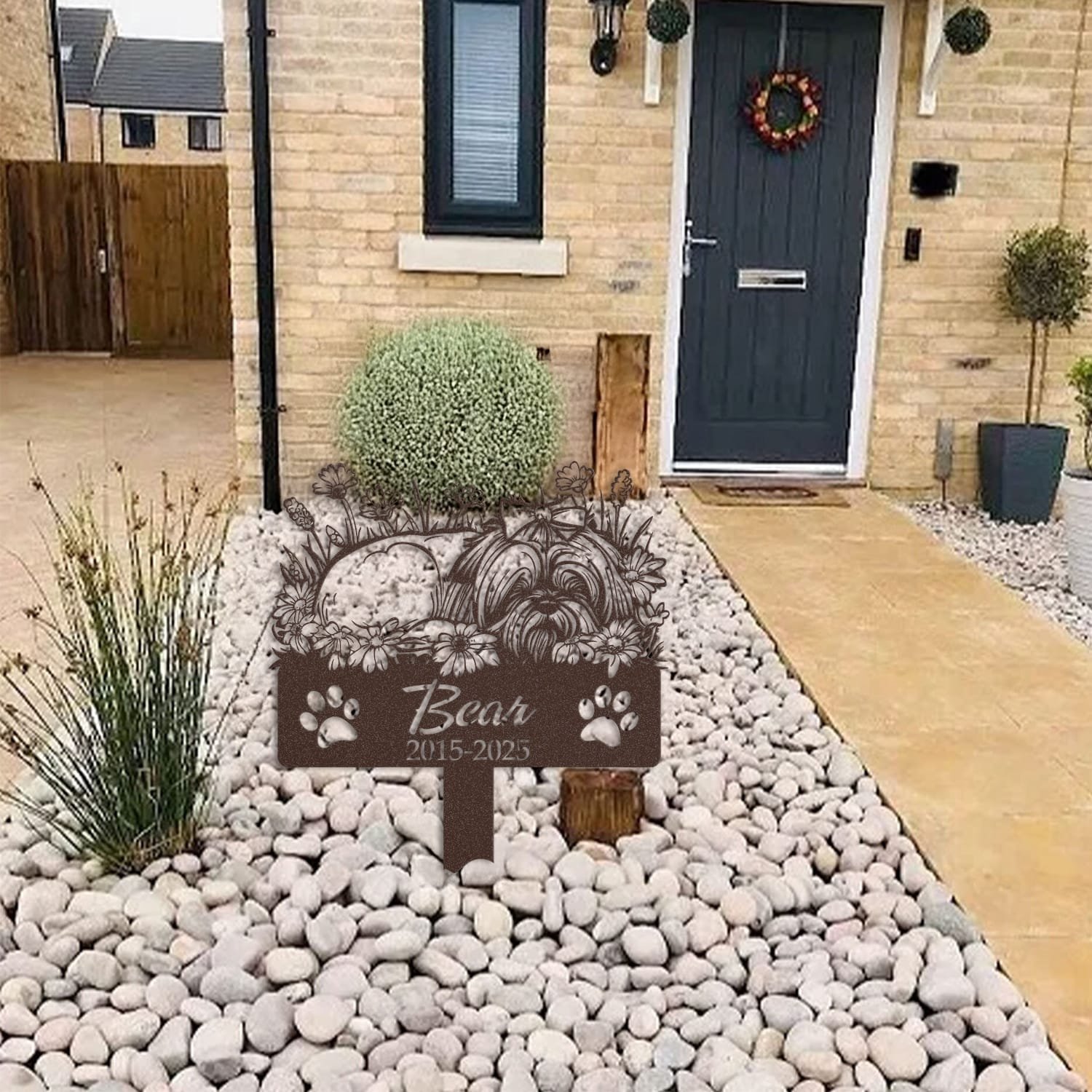 Long Hair Shih Tzu pet grave marker displayed among garden plants and stone edging, showing the sleeping-pose daisy floral design as an outdoor memorial accent.