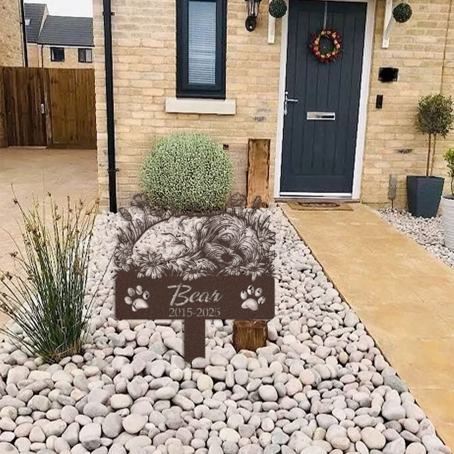 Maltese pet grave marker displayed among garden plants and stone edging, showing the sleeping-pose daisy floral design as an outdoor memorial accent.