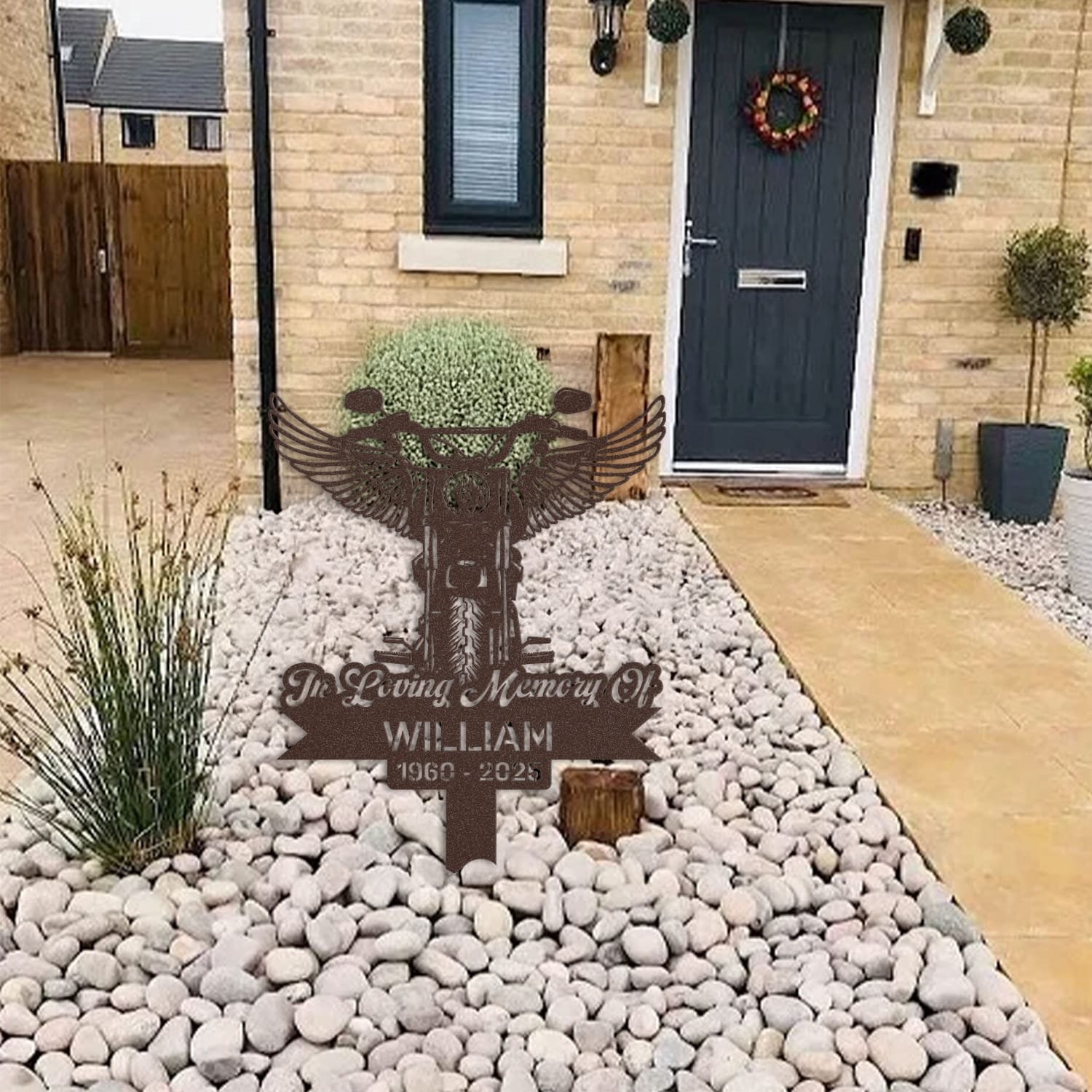 Motorcycle human grave marker displayed among garden plants and stone edging, showing the motorcycle angel wings design as an outdoor memorial accent.