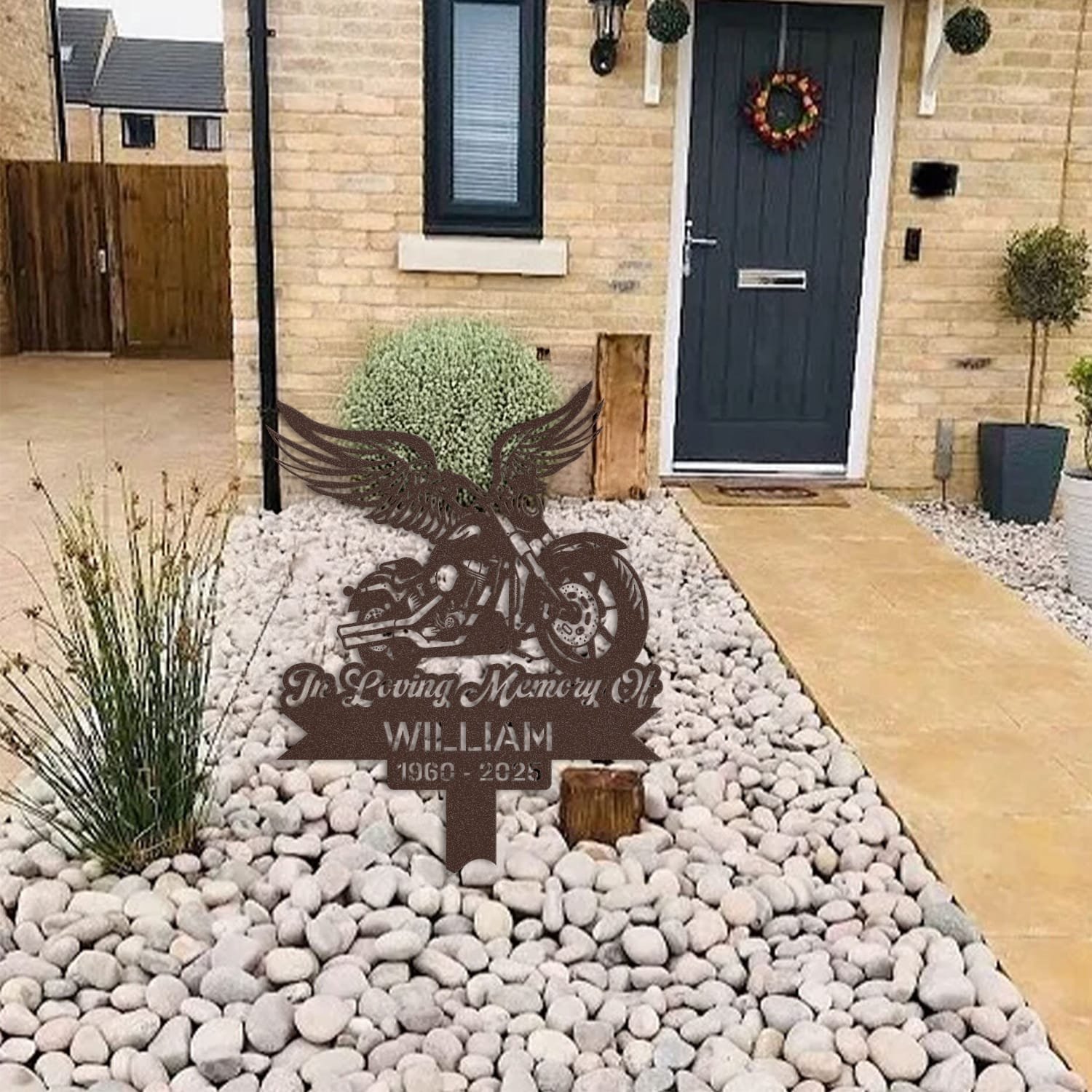 Motorcycle human grave marker displayed among garden plants and stone edging, showing the motorcycle winged rider design as an outdoor memorial accent.