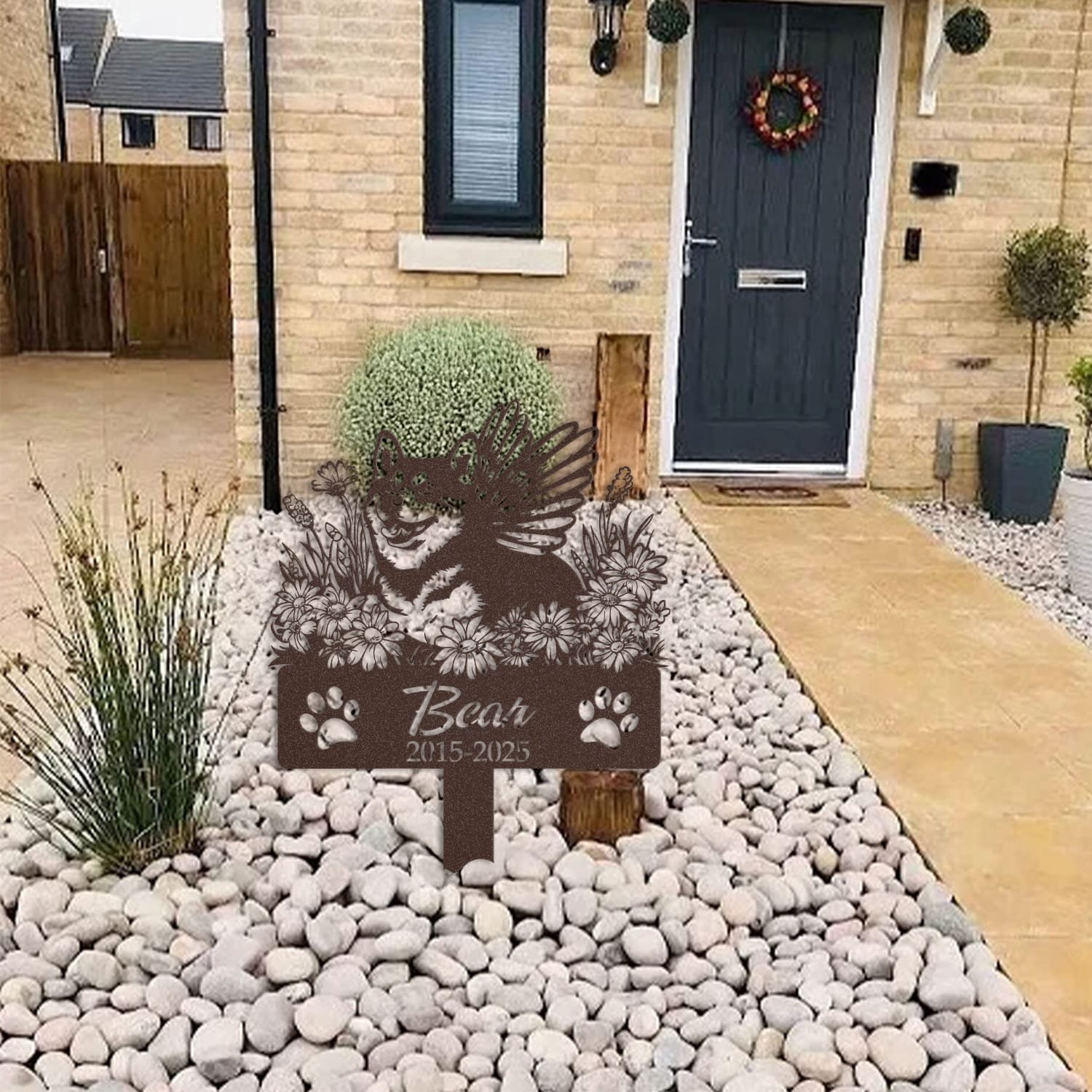 Shiba Inu pet grave marker displayed among garden plants and stone edging, showing the angel wings and daisy floral design as an outdoor memorial accent.
