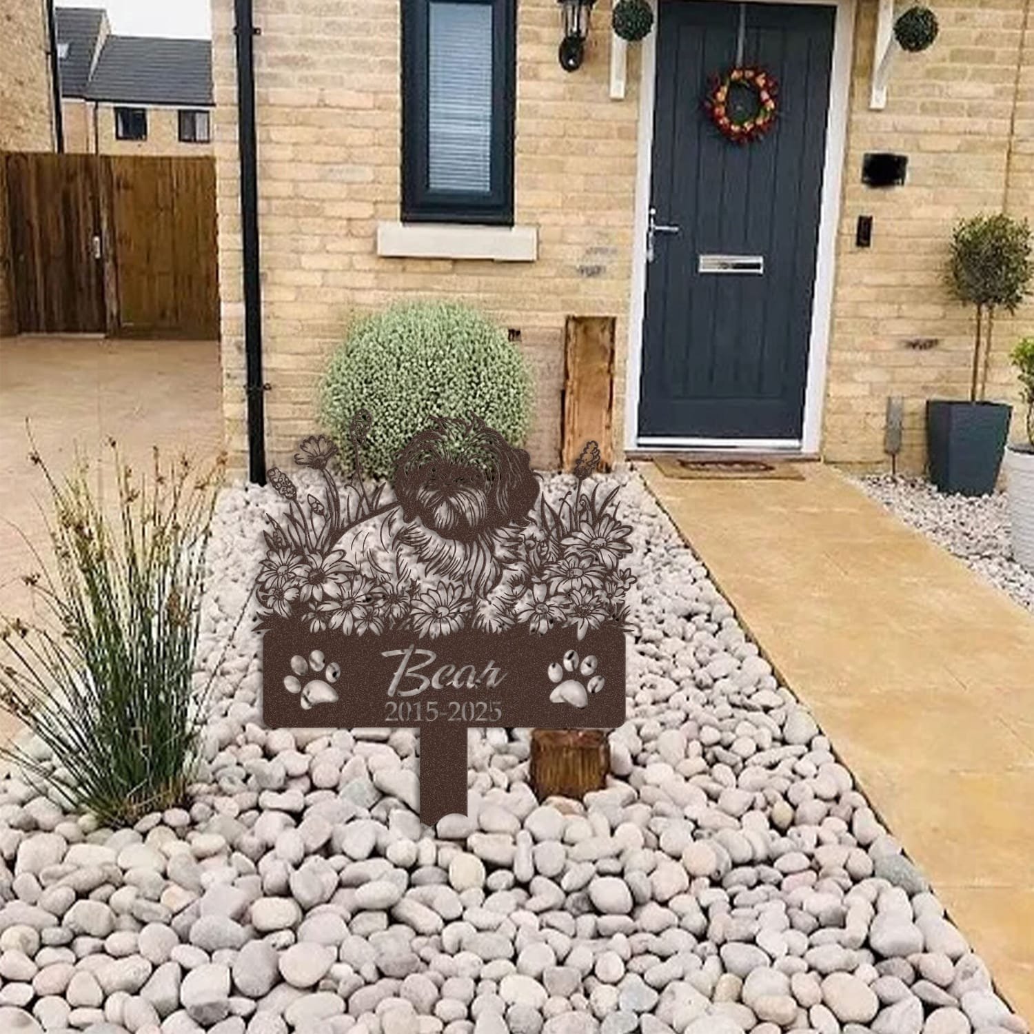 Shih Tzu pet grave marker displayed among garden plants and stone edging, showing the sleeping-pose daisy floral design as an outdoor memorial accent.