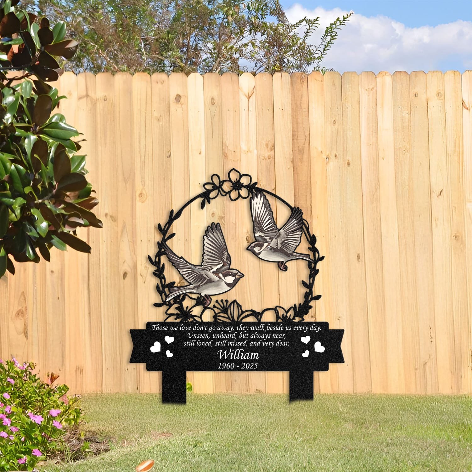 Sparrow human grave marker displayed among garden plants and stone edging, showing the floral wreath bird-pair design as an outdoor memorial accent.