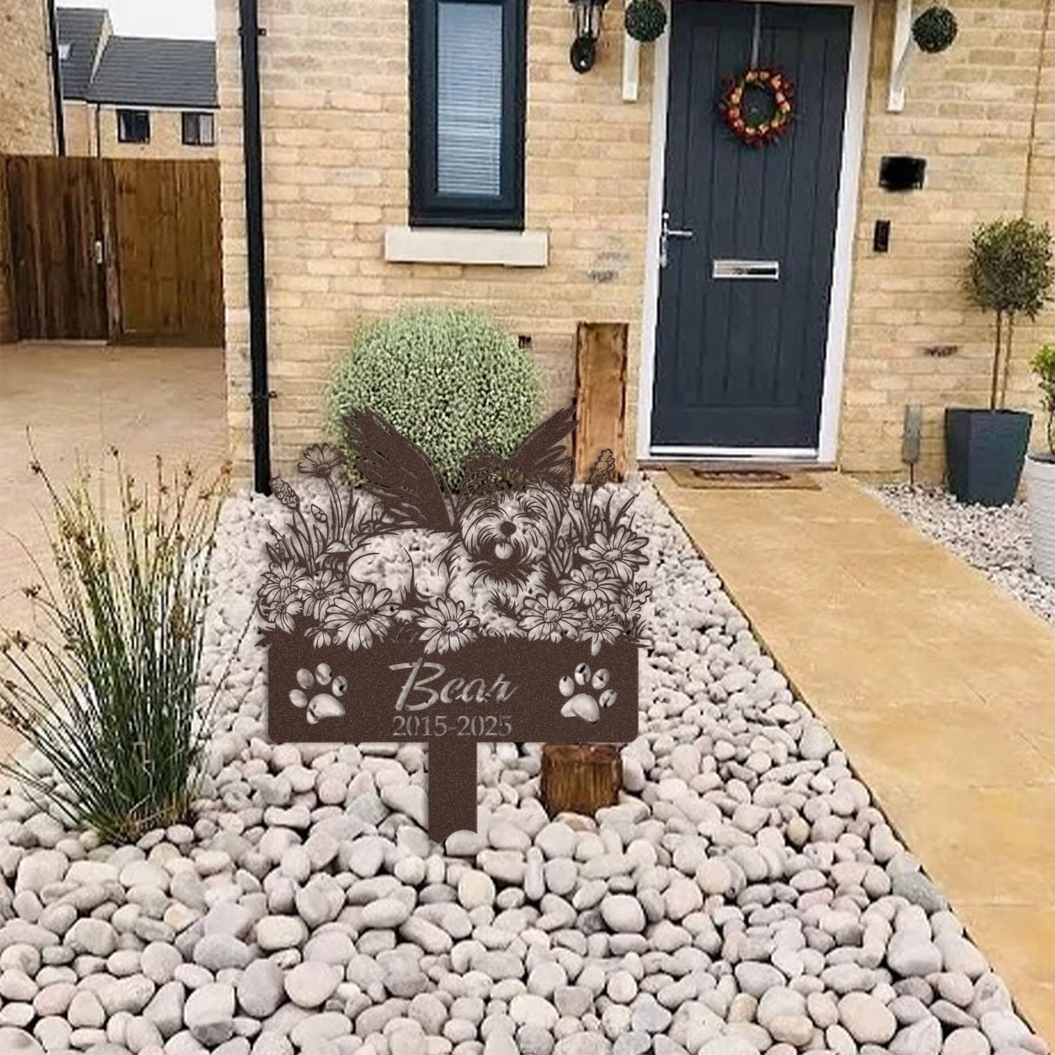 West Highland White Terrier pet grave marker displayed among garden plants and stone edging, showing the angel wings and daisy floral design as an outdoor memorial accent.
